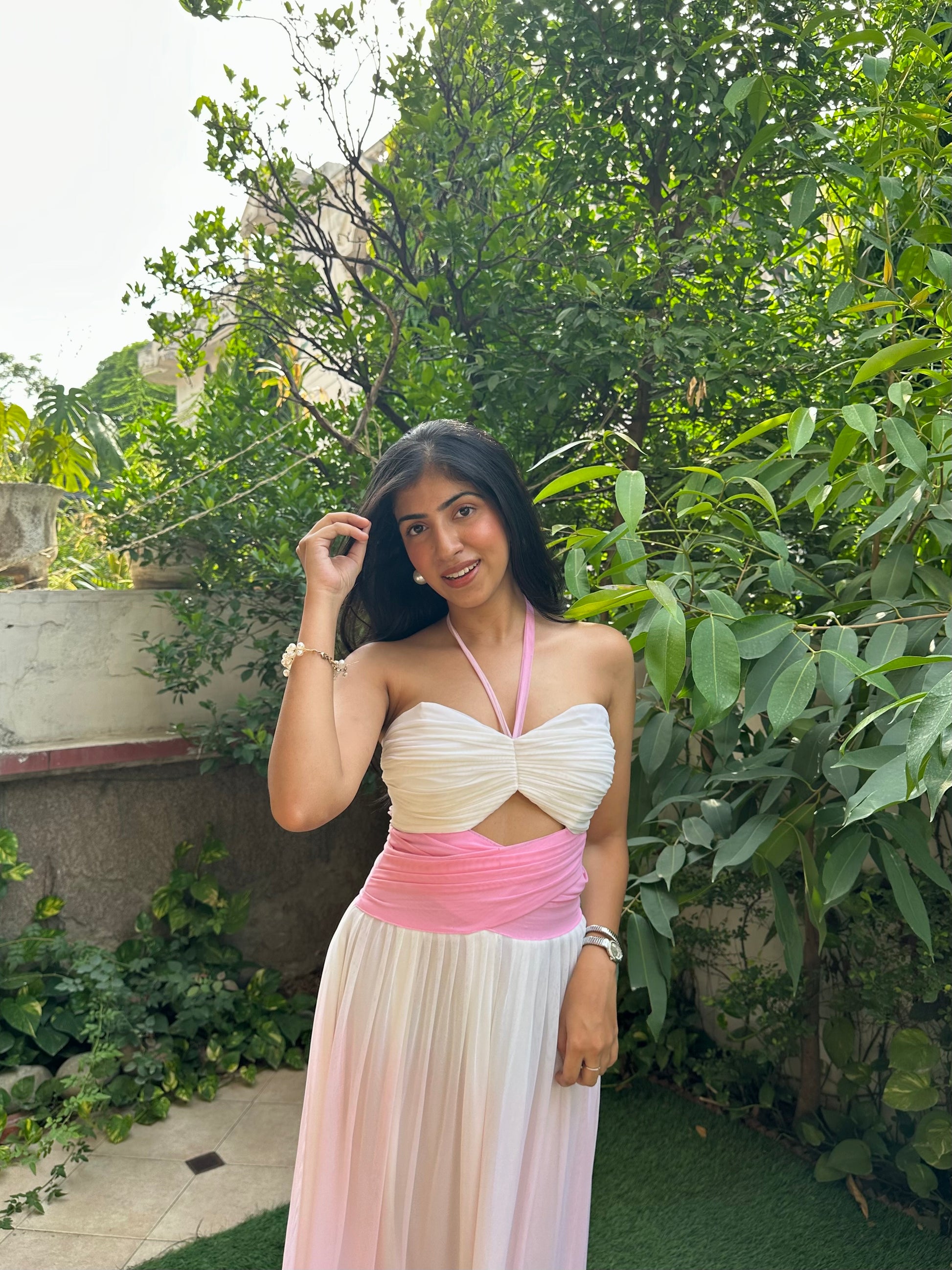 A woman posing outdoors in a sleeveless white and pink dress with a cutout at the waist, set against a background of green foliage and a stone wall.