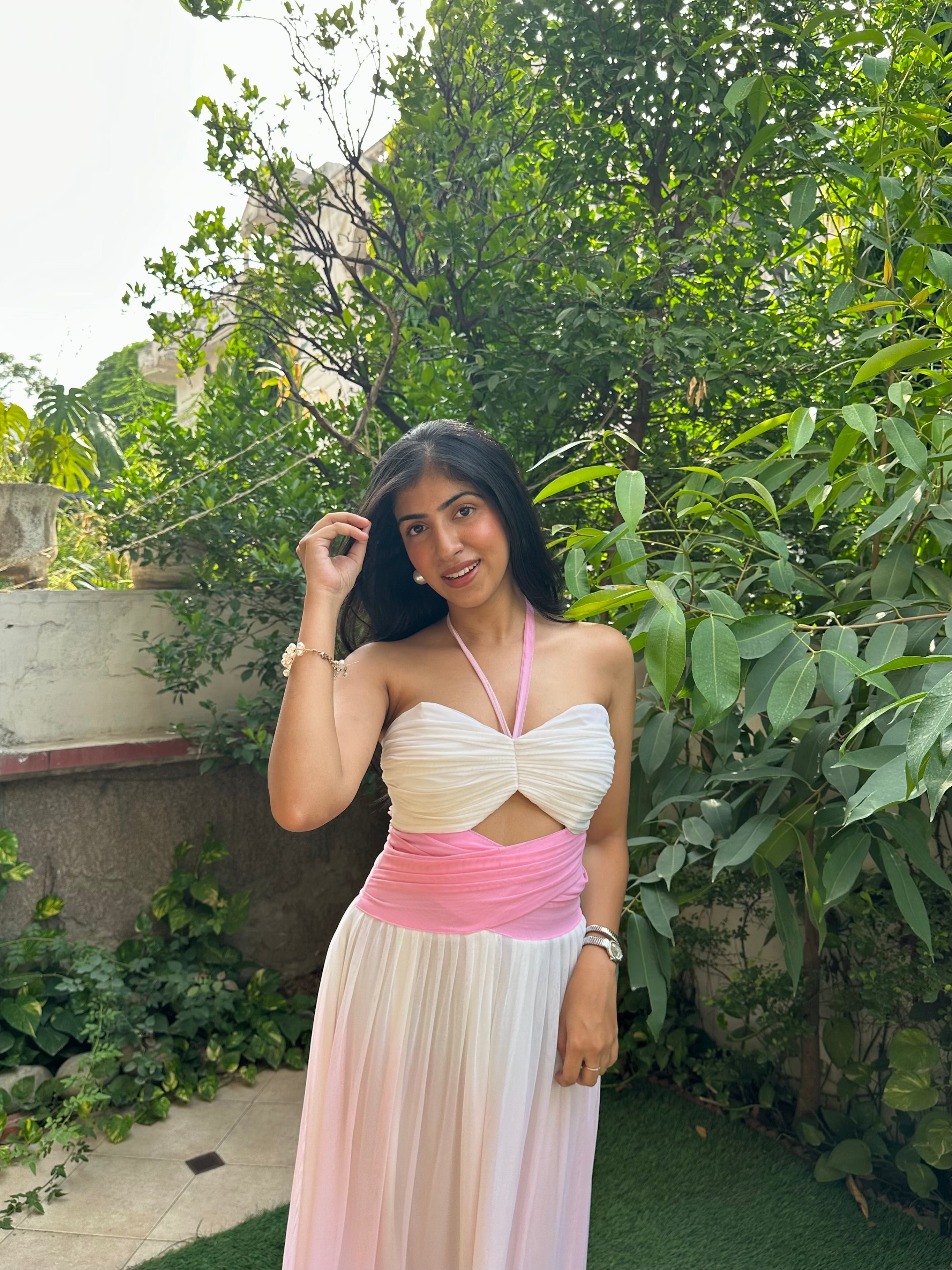 A woman posing outdoors in a sleeveless white and pink dress with a cutout at the waist, set against a background of green foliage and a stone wall.