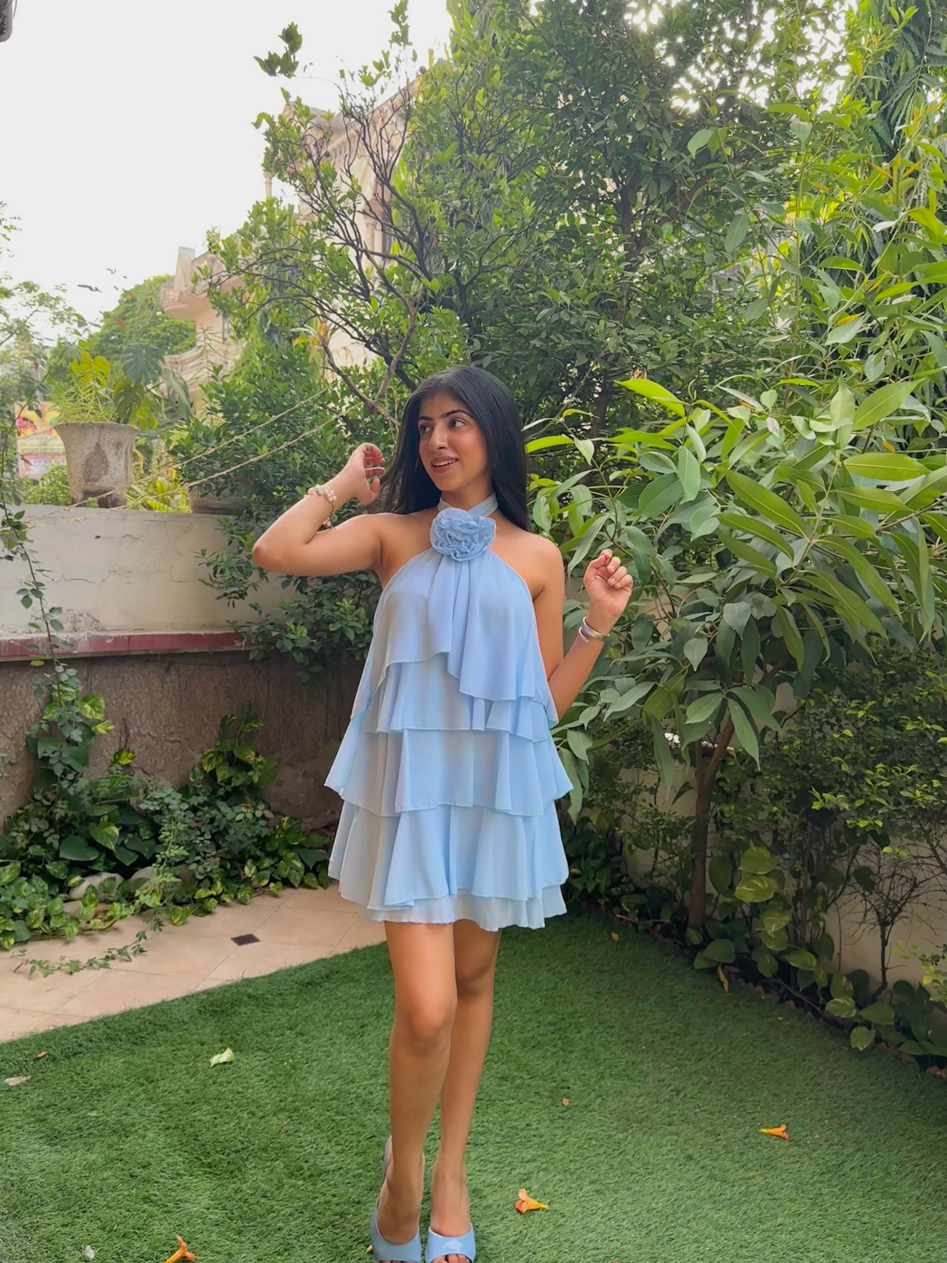 A woman wearing a light blue tiered halter mini dress with a large ruffled neckline, standing outdoors on green artificial grass with leafy plants and a wall in the background.
