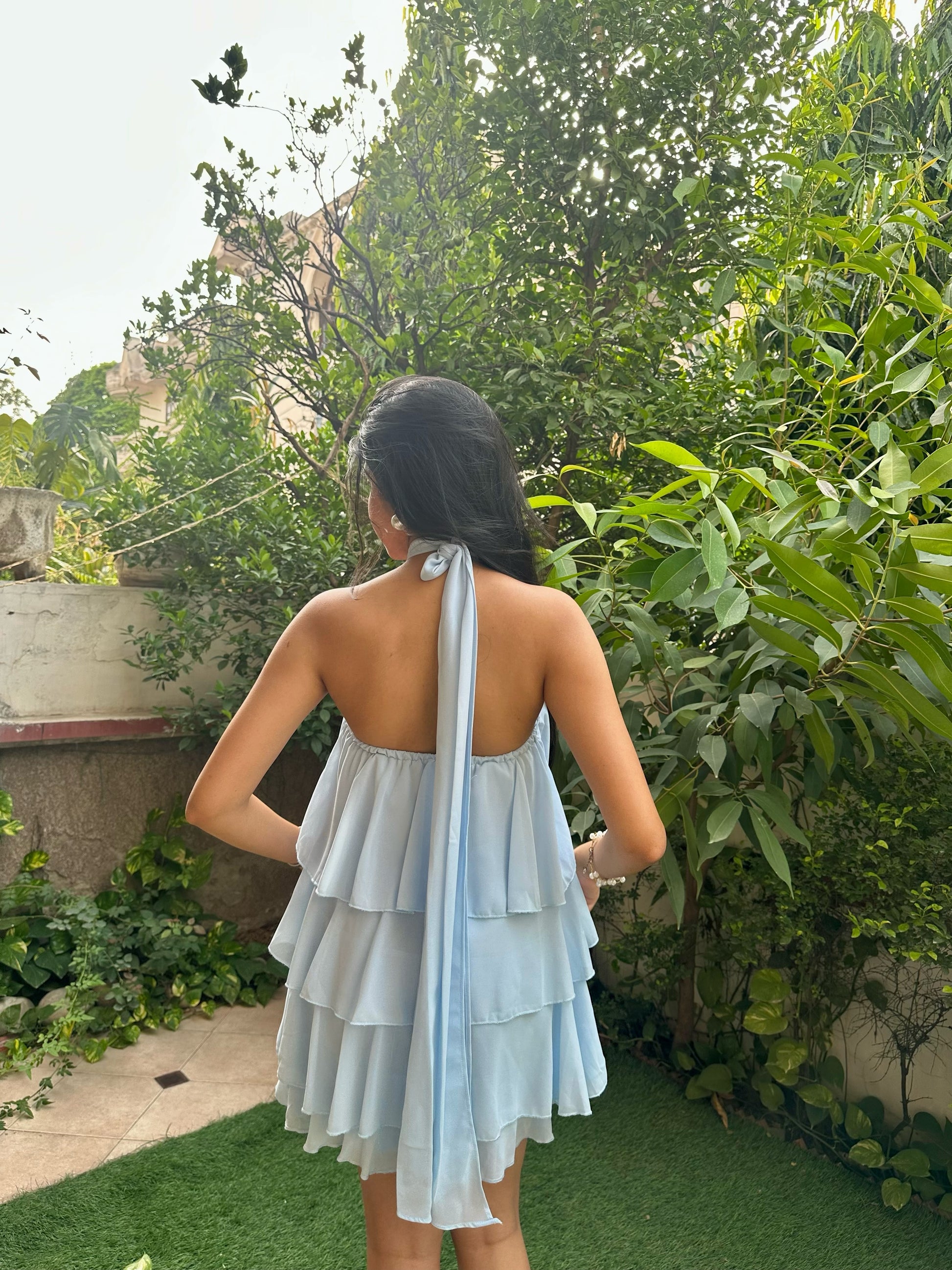 A woman viewed from behind wearing a light blue halter mini dress with a tiered ruffle skirt and a long tie-back ribbon, standing in a garden setting with green foliage.